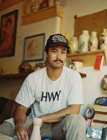 A man with a mustache and a baseball cap sits in a pottery studio. He is wearing a t-shirt and is holding a ceramic vase.