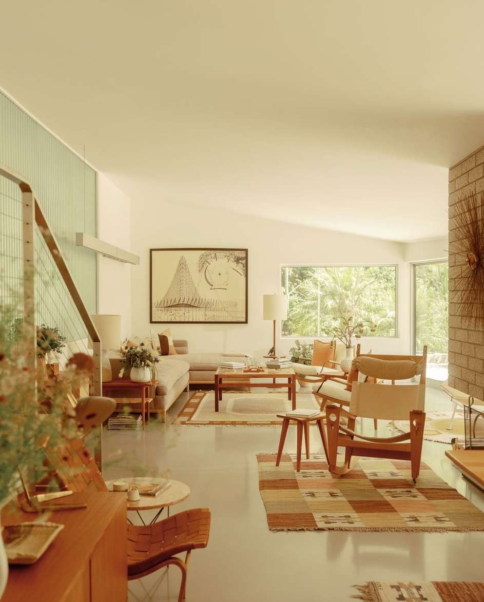 Mid-century modern living room interior with neutral palette, wood furniture, area rugs, brick fireplace, and leafy green outdoor view.