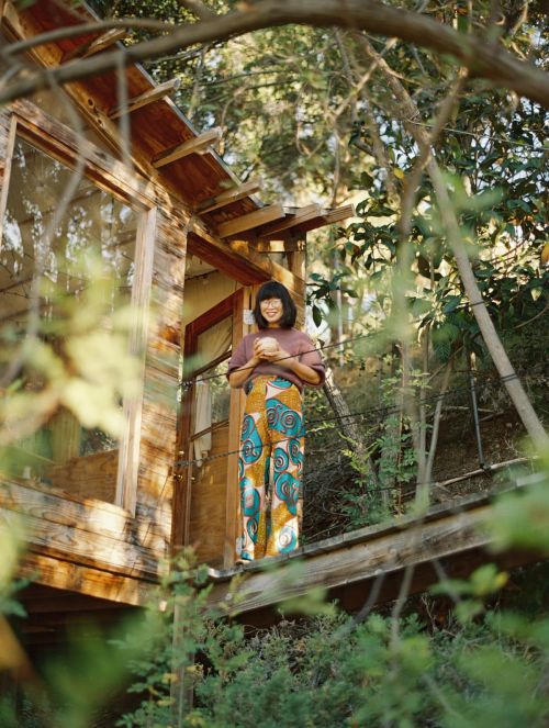 Woman in patterned pants stands on a wood deck surrounded by lush green foliage, holding a round object, with a partially visible house.