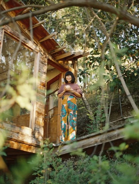 Woman in patterned pants stands on a wood deck surrounded by lush green foliage, holding a round object, with a partially visible house.