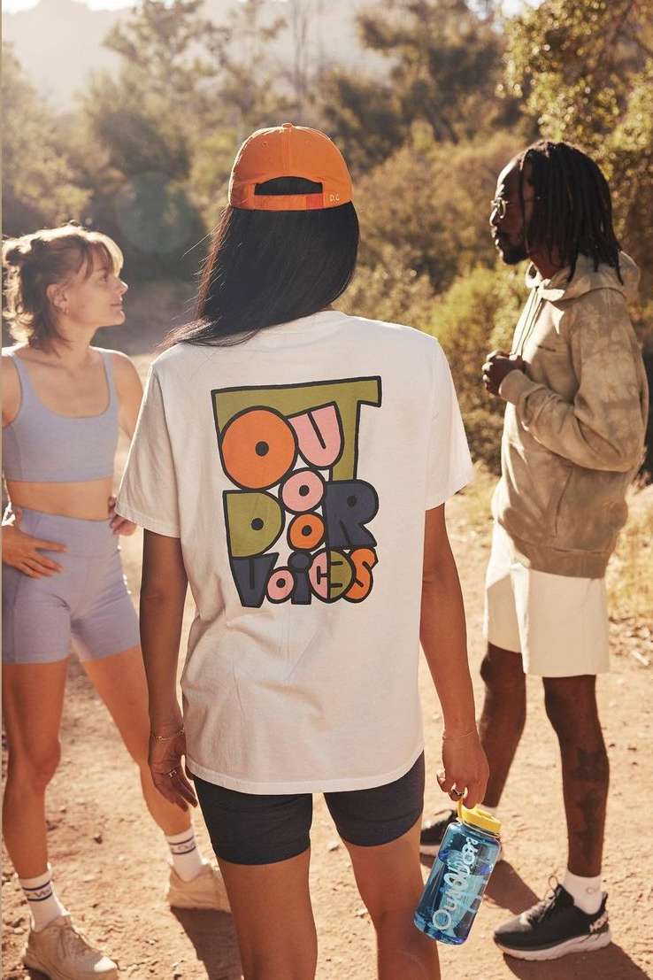 Three people on a dirt path, one woman in the foreground wearing a white T-shirt with a graphic and an orange baseball cap.