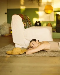 Woman resting her head on her arms on a woven rug next to a white armchair and straw hat