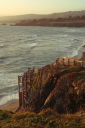 Elevated coastal view of wooden stairs descending a rocky cliff toward a sandy beach and ocean at sunset.