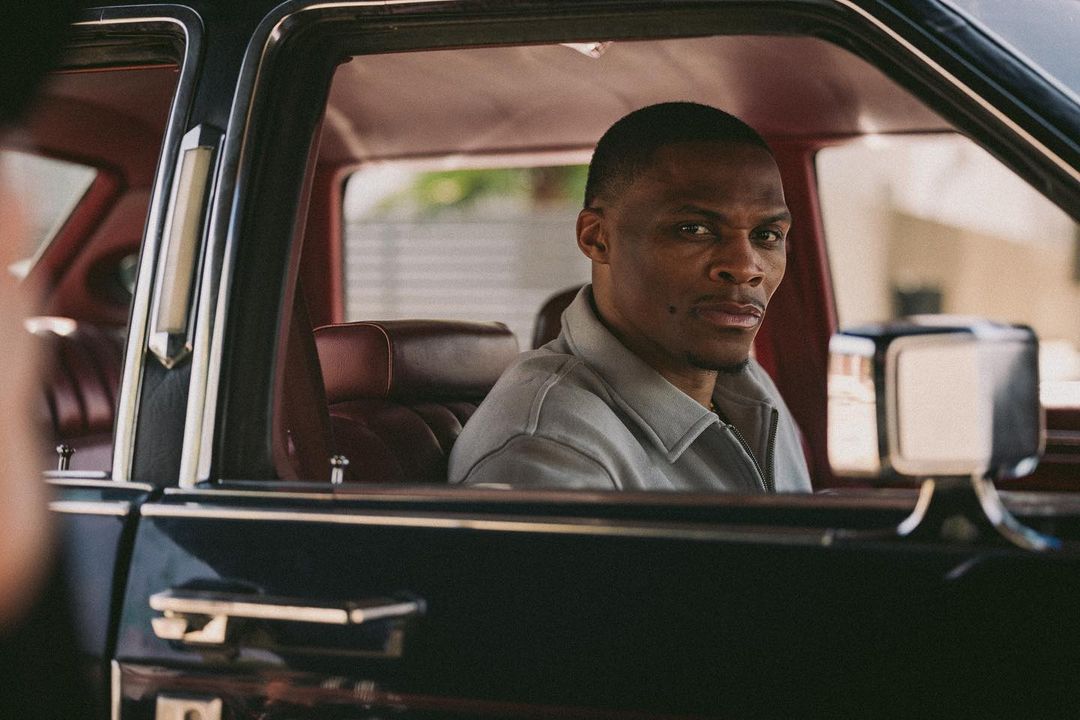 An adult male sits in the driver's seat of a black luxury car, looking towards the viewer. Red leather interior and car details visible.