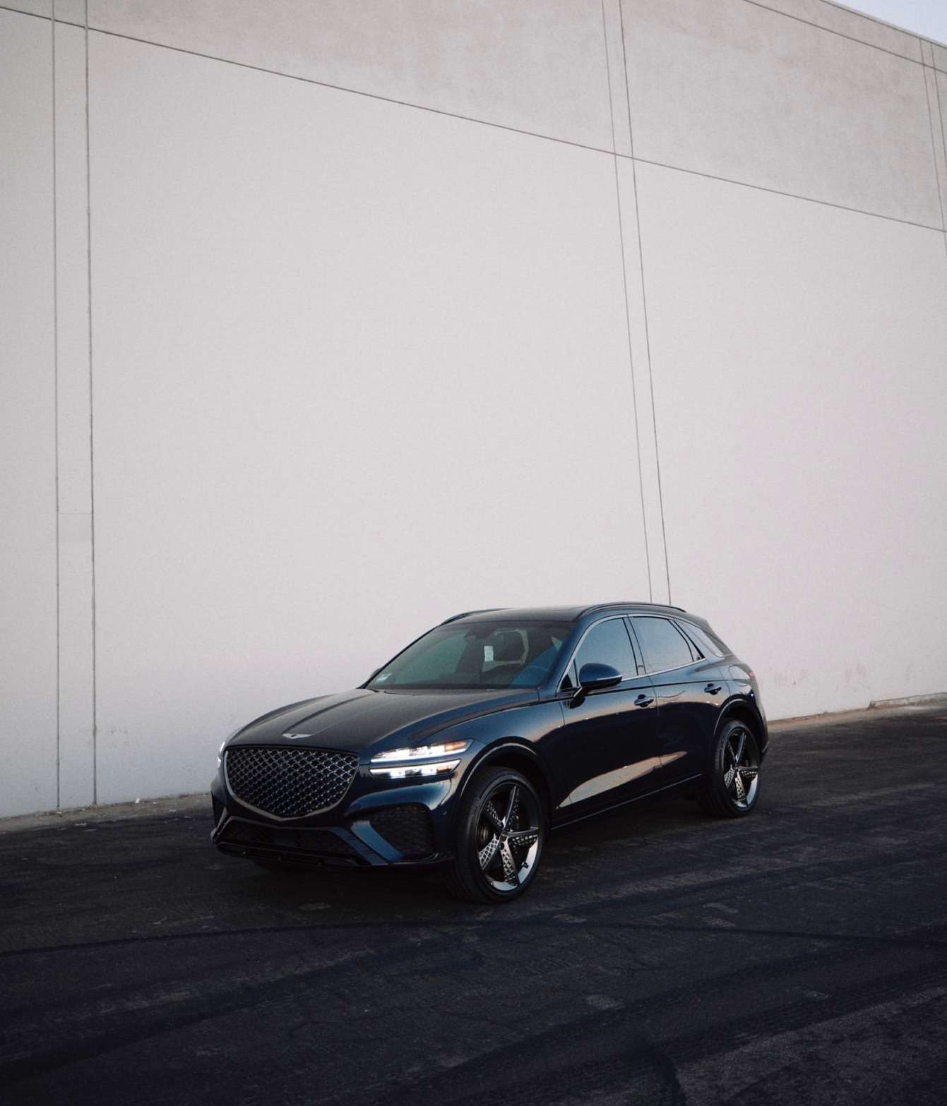 Dark blue Genesis GV70 SUV parked against a tall, light gray wall on dark asphalt, showcasing its sleek design and modern aesthetic.