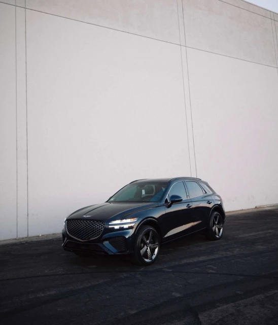 Dark blue Genesis GV70 SUV parked against a tall, light gray wall on dark asphalt, showcasing its sleek design and modern aesthetic.