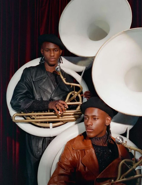 Two adult male musicians wearing black berets and leather jackets posing with tubas in front of a red curtain in an indoor setting.