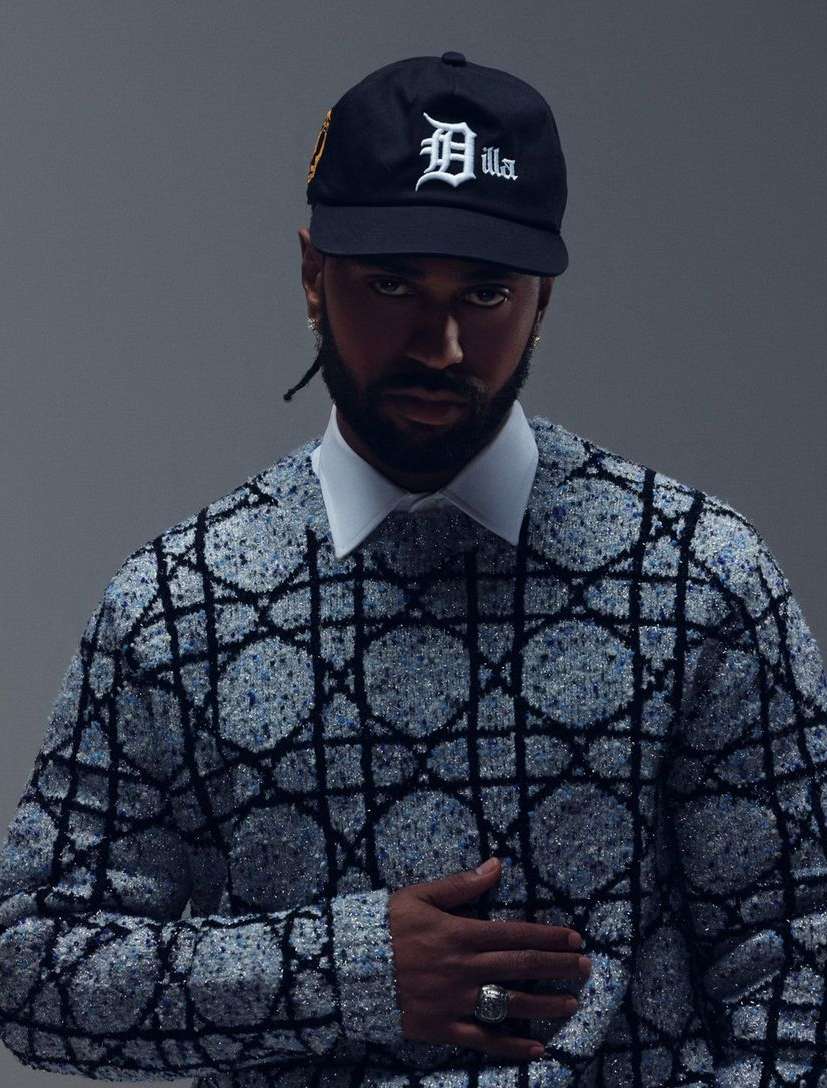Adult male poses indoors wearing a baseball cap, patterned sweater and collared shirt against a gray background in a medium shot.