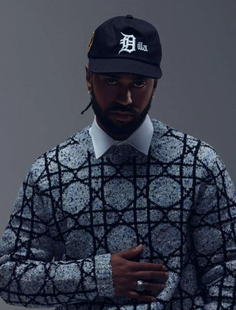 Adult male poses indoors wearing a baseball cap, patterned sweater and collared shirt against a gray background in a medium shot.