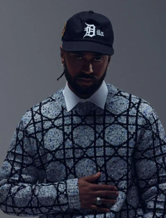 Adult male poses indoors wearing a baseball cap, patterned sweater and collared shirt against a gray background in a medium shot.