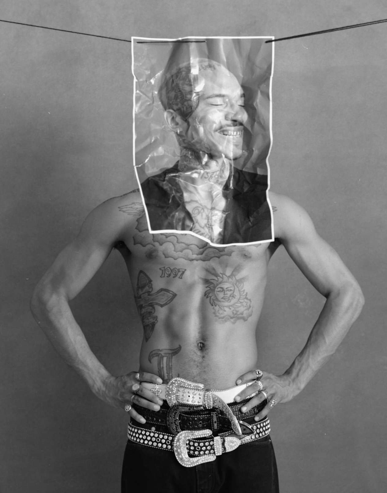 Black and white studio shot of a shirtless man with tattoos, wearing several studded belts, and holding a picture of his face.