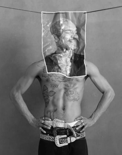 Black and white studio shot of a shirtless man with tattoos, wearing several studded belts, and holding a picture of his face.