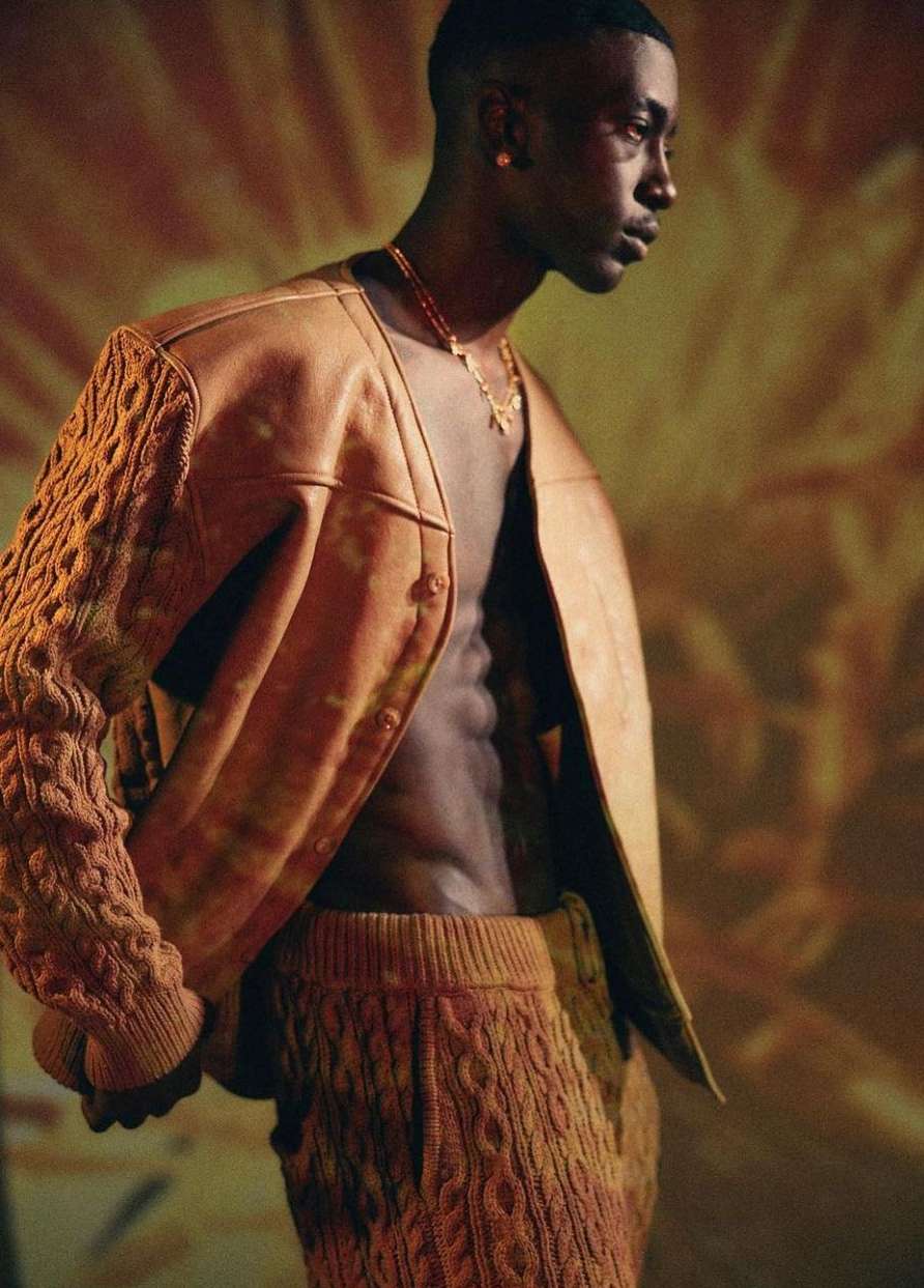 Adult man wearing an open leather cardigan with knitted sleeves and matching knitted pants poses against a diffused warm toned background.