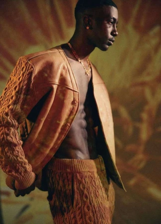 Adult man wearing an open leather cardigan with knitted sleeves and matching knitted pants poses against a diffused warm toned background.