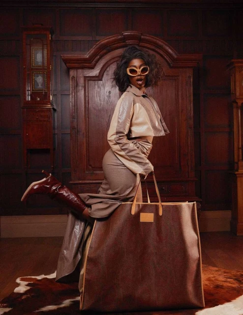 A Black woman in a beige outfit poses indoors with a very large brown handbag, set against a paneled wood wall with vintage furniture.