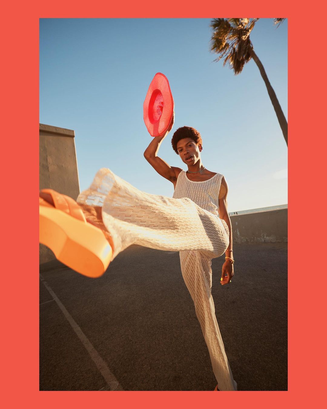 An androgynous adult with a pink hat poses with one leg raised and a wide yellow platform shoe; a palm tree accents the skyline.