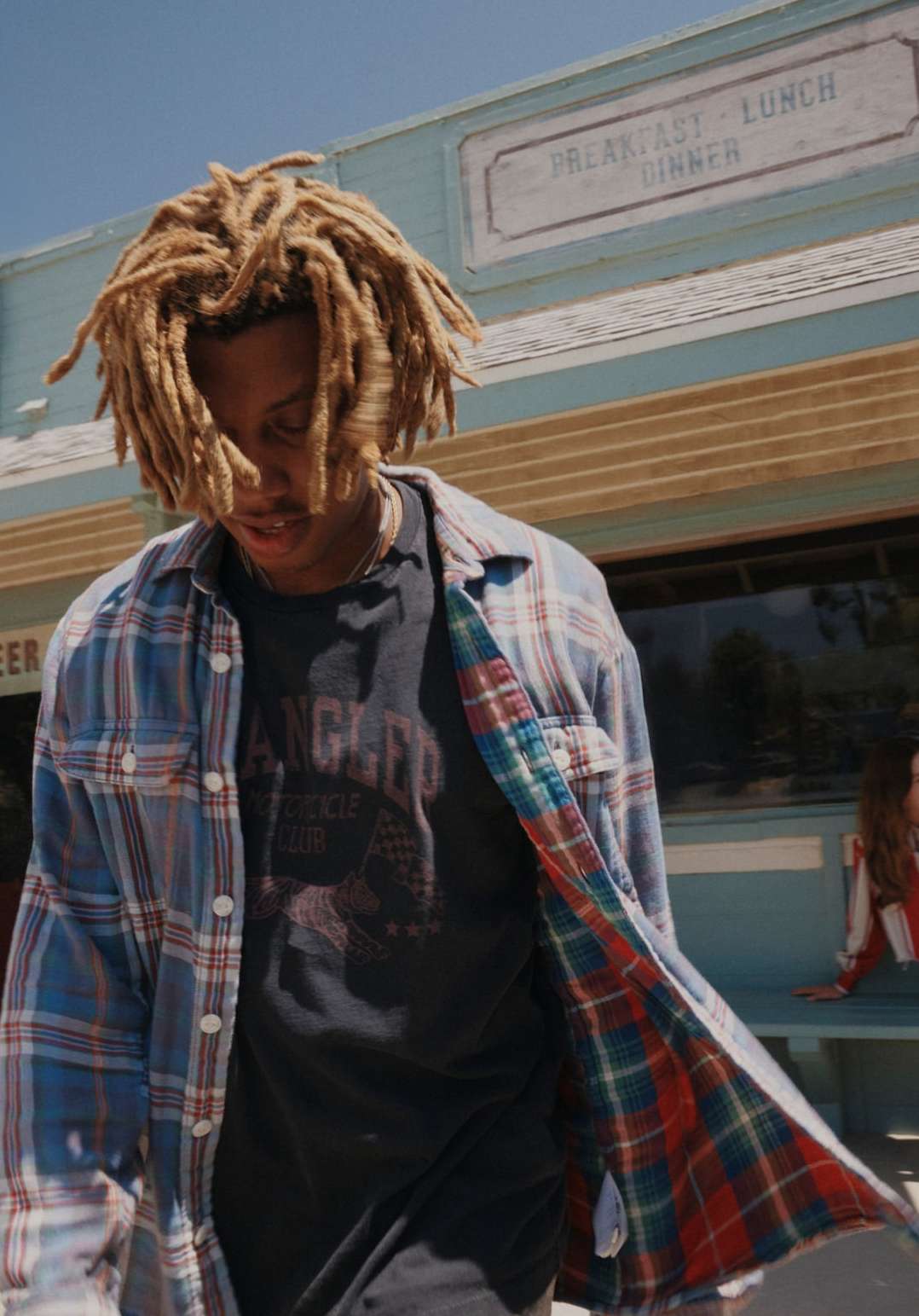 A young adult with dreadlocks wears an open flannel shirt over a t-shirt outdoors in front of a diner in this eye-level medium shot.
