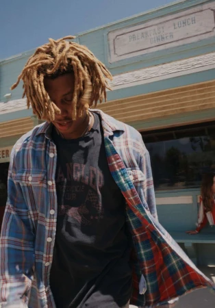 A young adult with dreadlocks wears an open flannel shirt over a t-shirt outdoors in front of a diner in this eye-level medium shot.