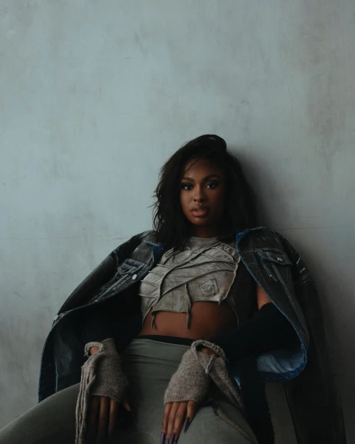Portrait of an African-American woman in a grey crop top and a dark blue denim jacket posing against a textured grey wall.