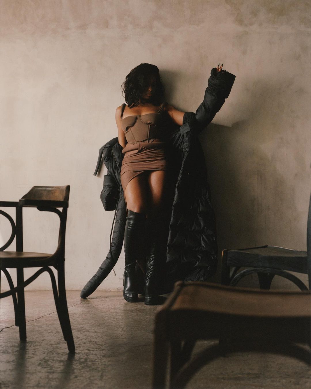 Brunette woman poses in a brown corset dress and black boots with a puffy black jacket in a sparse indoor environment with chairs.