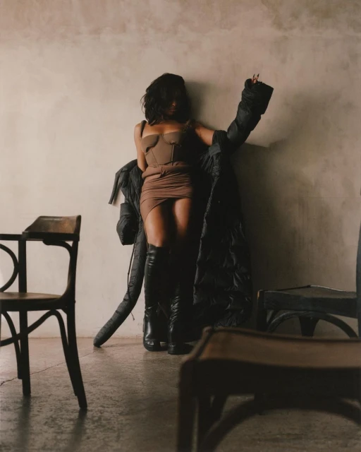 Brunette woman poses in a brown corset dress and black boots with a puffy black jacket in a sparse indoor environment with chairs.