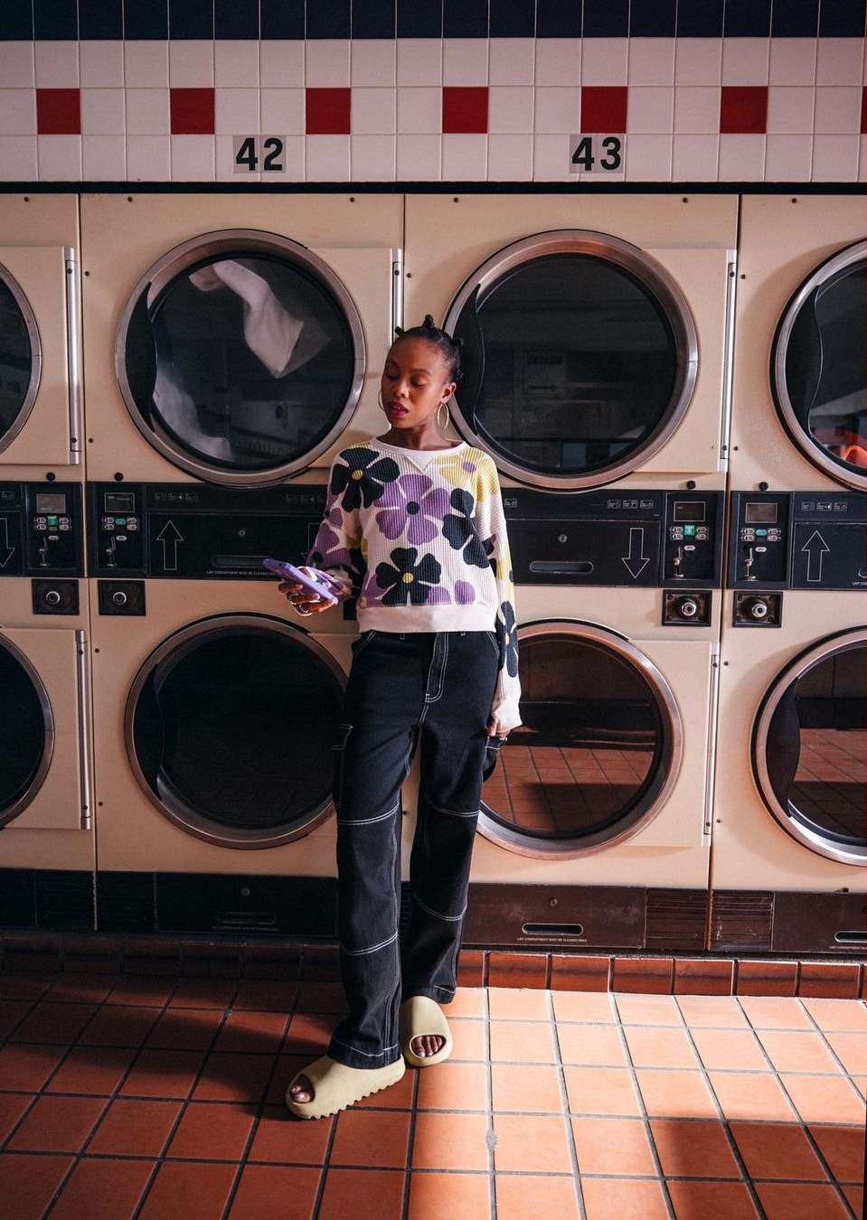 Woman in floral sweater and black pants poses in front of rows of washing machines in a brightly lit laundromat setting.
