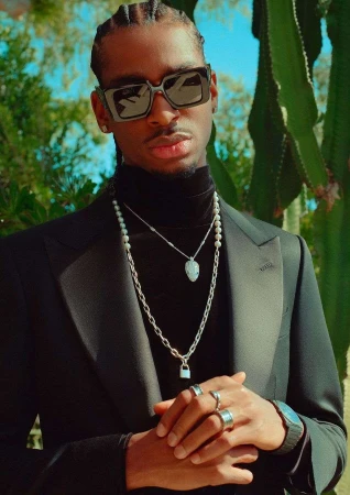 Portrait of a black man wearing sunglasses, black suit jacket, turtleneck, layered silver chains, and rings outdoors against cacti