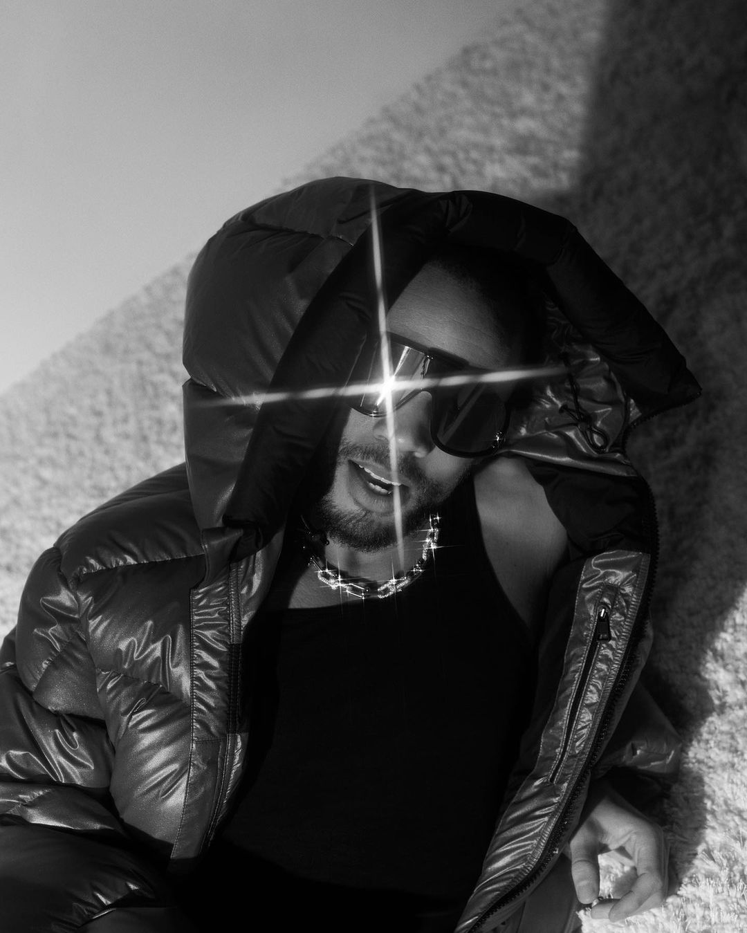 Black and white close-up of a male model in sunglasses and a puffy jacket, reclining against a soft, textured surface indoors.