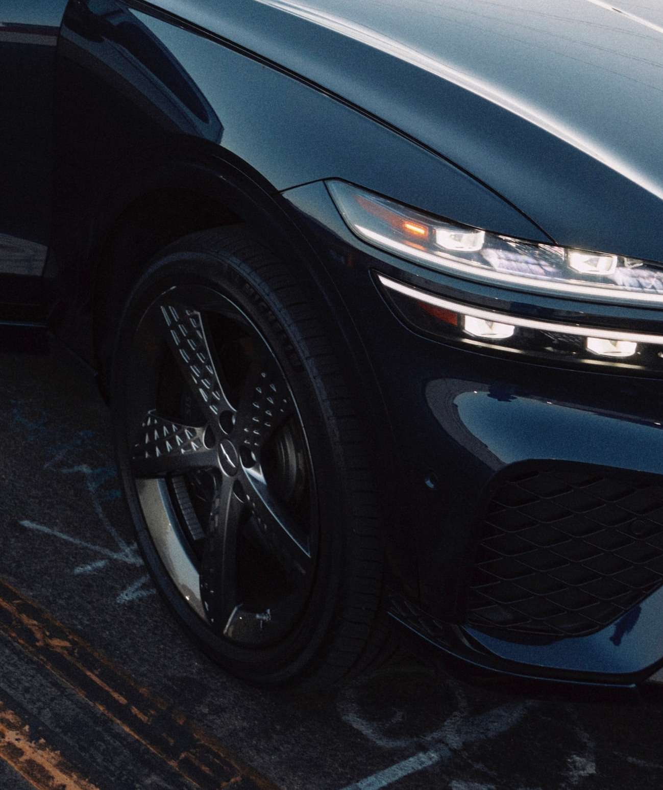 Close-up shot of a dark blue Genesis GV80 SUV showcasing its sleek headlights, front-end design, and patterned rims on the front wheel.