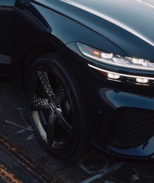 Close-up shot of a dark blue Genesis GV80 SUV showcasing its sleek headlights, front-end design, and patterned rims on the front wheel.