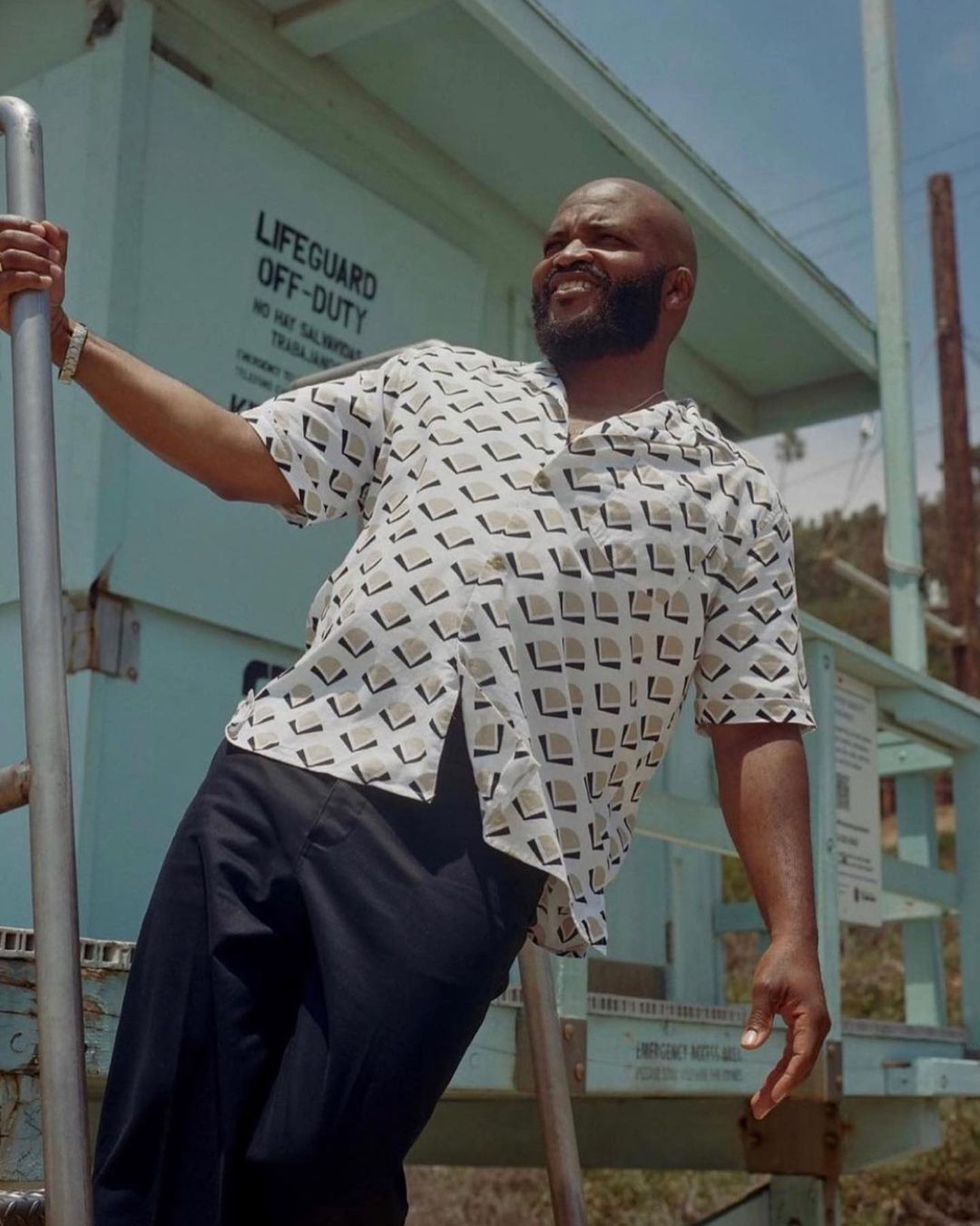 An African American man smiles broadly on a lifeguard tower with a geometric patterned shirt, exuding a relaxed, casual style outdoors.