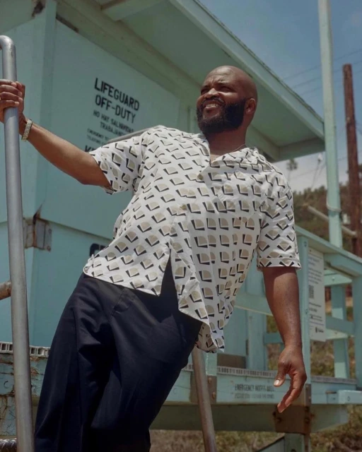 An African American man smiles broadly on a lifeguard tower with a geometric patterned shirt, exuding a relaxed, casual style outdoors.