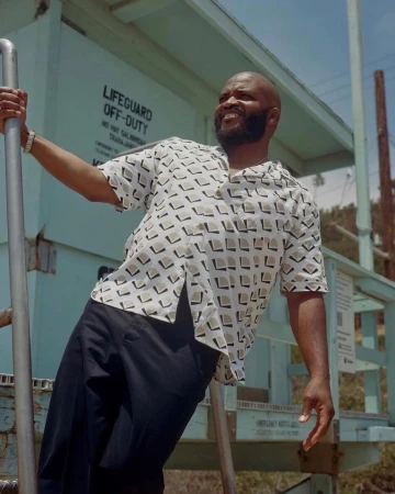 An African American man smiles broadly on a lifeguard tower with a geometric patterned shirt, exuding a relaxed, casual style outdoors.