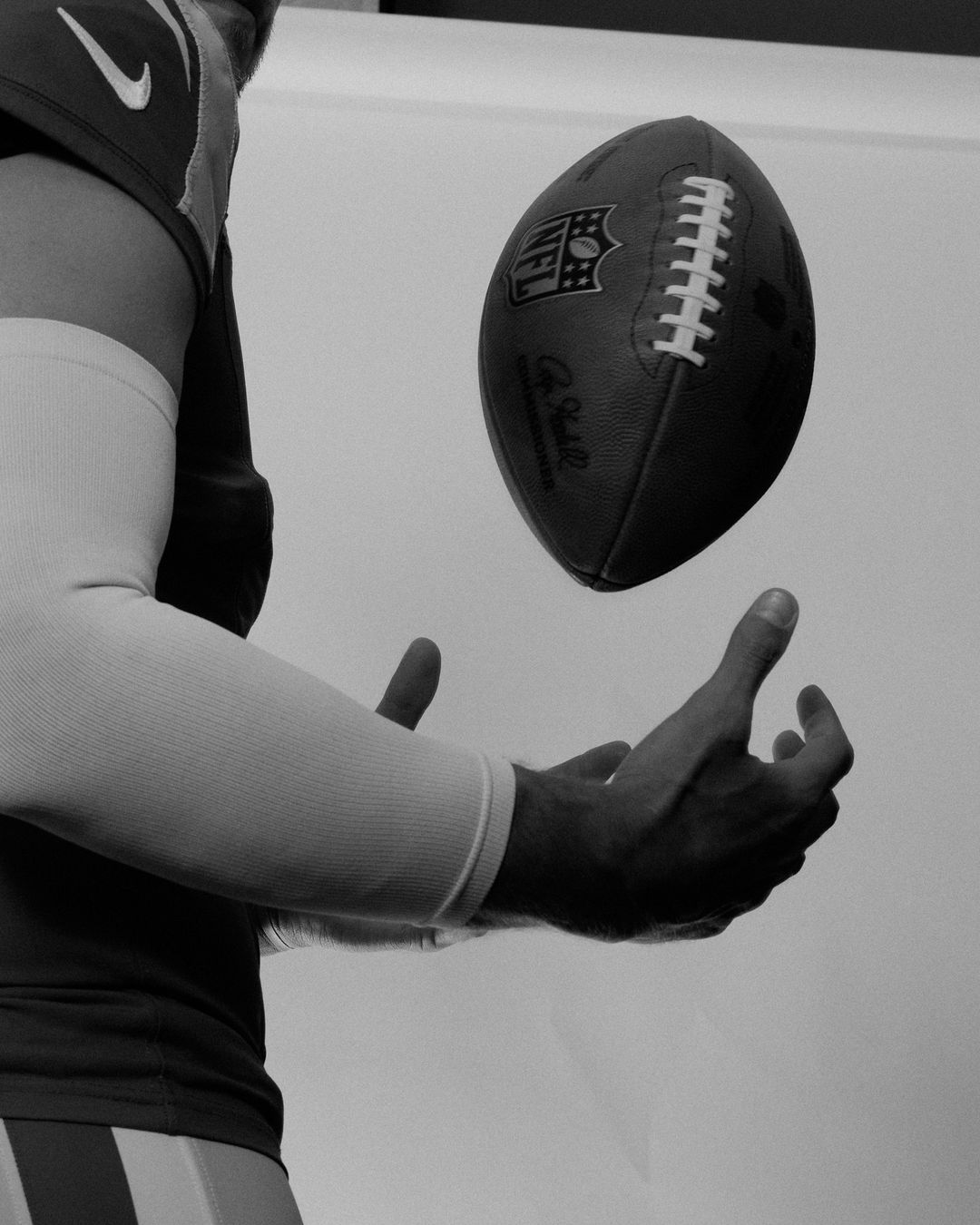 Monochrome image of an American football suspended over a person's hands and forearm, in front of a plain background.