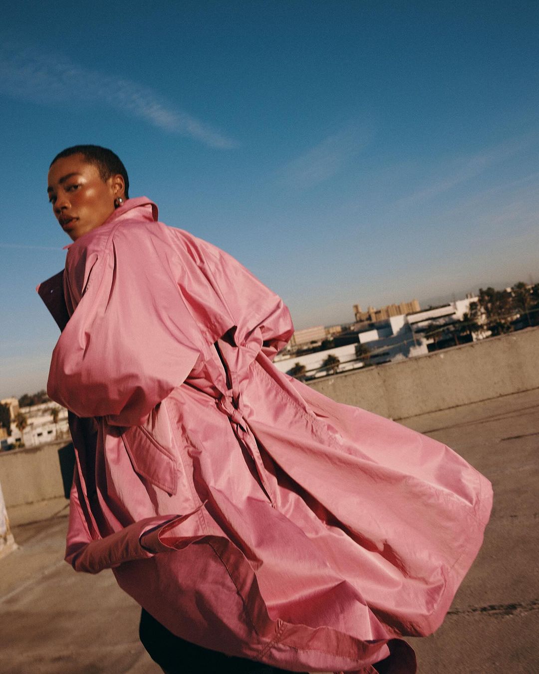 Model with short hair wearing a voluminous pink coat standing on a concrete rooftop against a clear blue sky