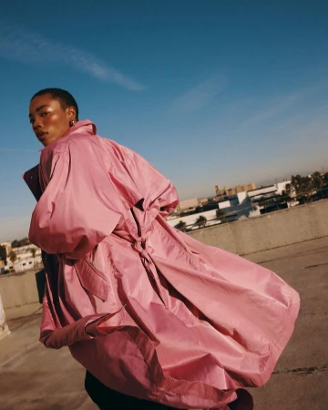 Model with short hair wearing a voluminous pink coat standing on a concrete rooftop against a clear blue sky