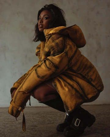 Black woman crouching wearing a mustard yellow jacket and black boots against a textured concrete wall