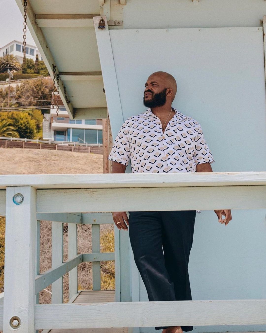 Adult male stands in a lifeguard tower on a sunny day, wearing a patterned shirt and dark pants with a beach and houses visible behind.