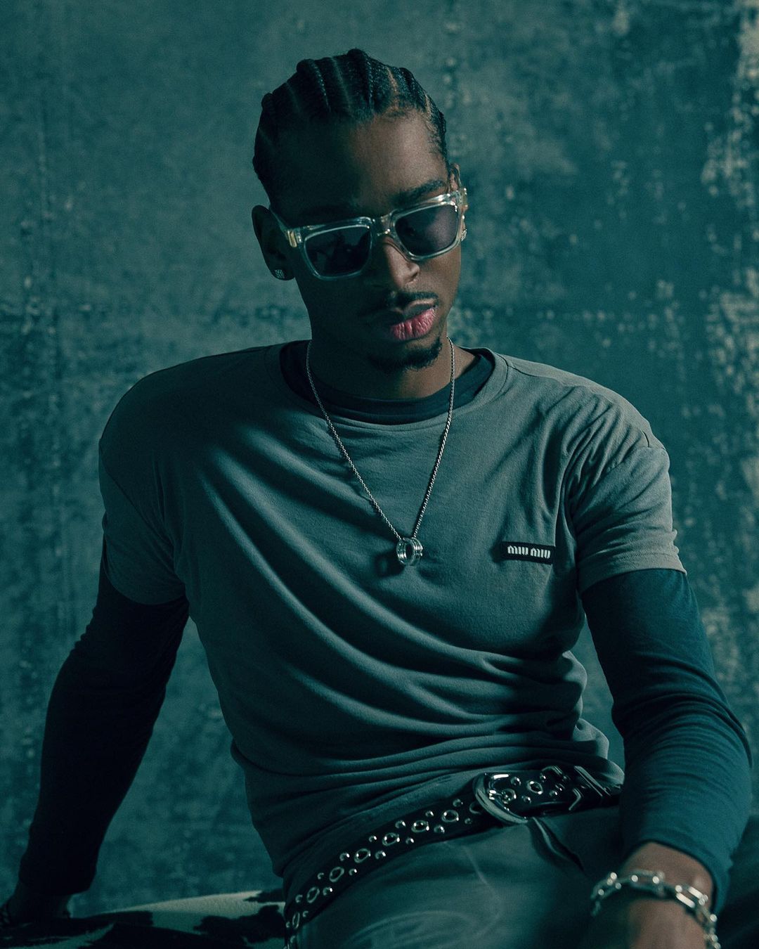 Portrait of a Black man with braids wearing sunglasses, a grey shirt, and silver jewellery, set against a textured blue background.