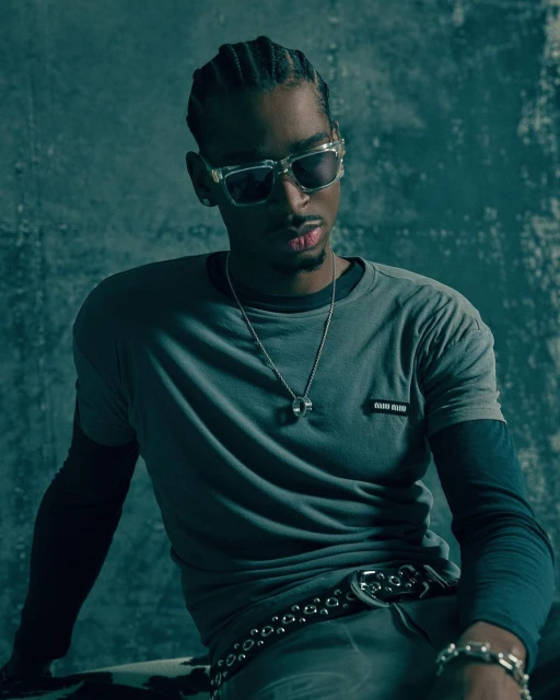 Portrait of a Black man with braids wearing sunglasses, a grey shirt, and silver jewellery, set against a textured blue background.