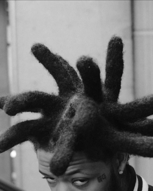 Close-up black and white portrait of a Black man with a serious expression, distinct dreadlocks, and visible facial tattoos.