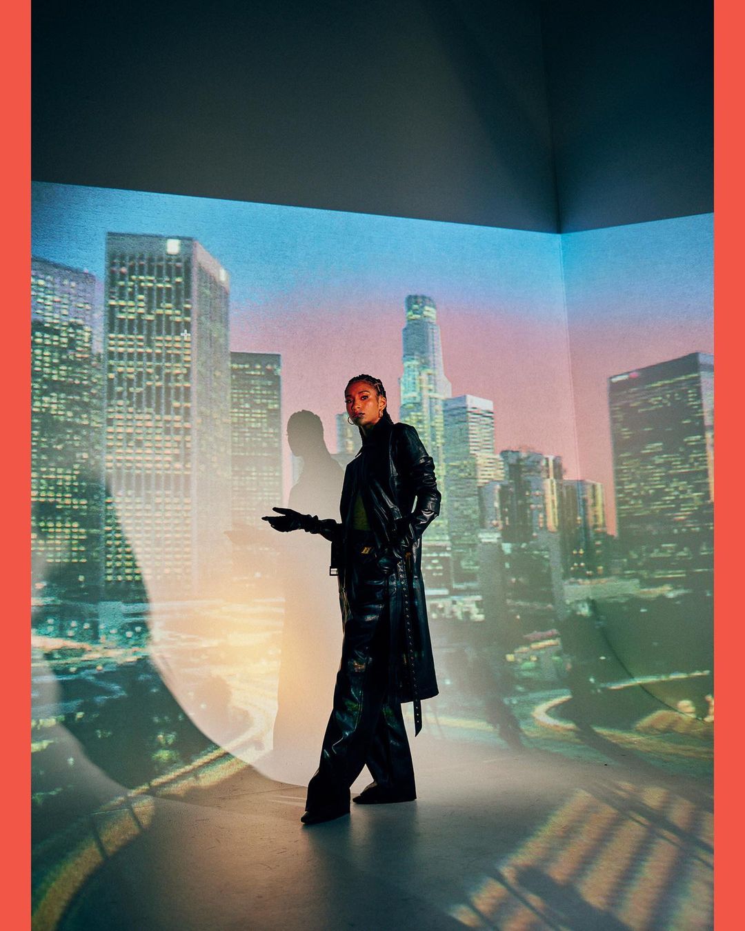 Person in black outfit stands in front of a cityscape projected onto a wall, with a spotlight creating a soft, diffused look.