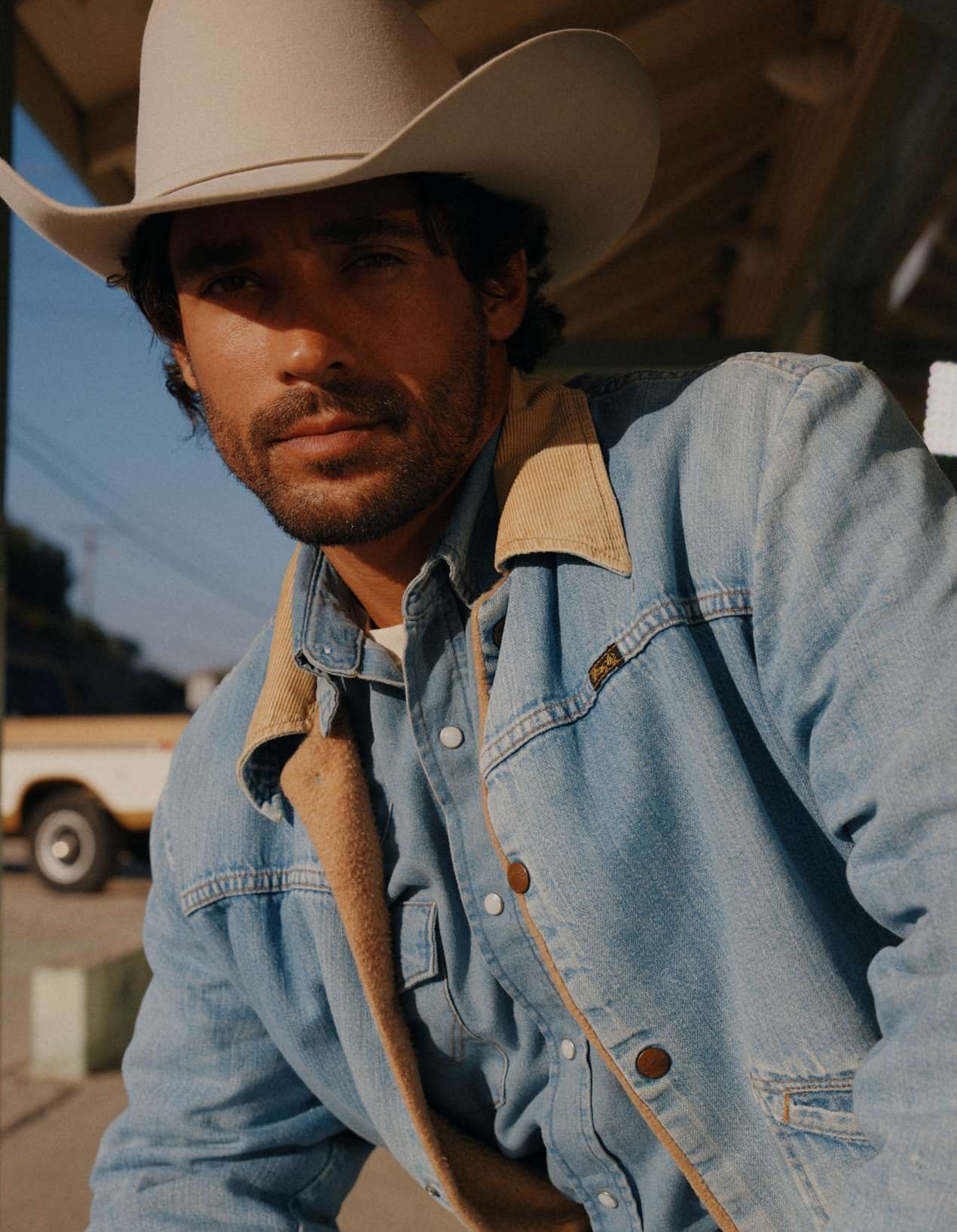 Close-up of a man in a cowboy hat and denim jacket, staring directly at the camera, with a vintage yellow truck in the background.