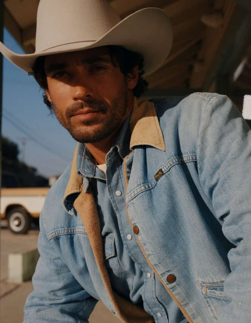 Close-up of a man in a cowboy hat and denim jacket, staring directly at the camera, with a vintage yellow truck in the background.