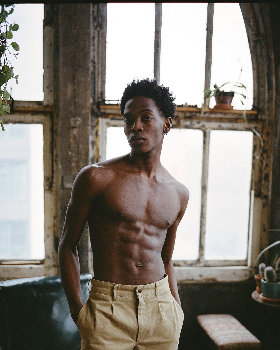 Portrait of shirtless African-American man in tan pants posing indoors near large windows with peeling paint and hanging plants.