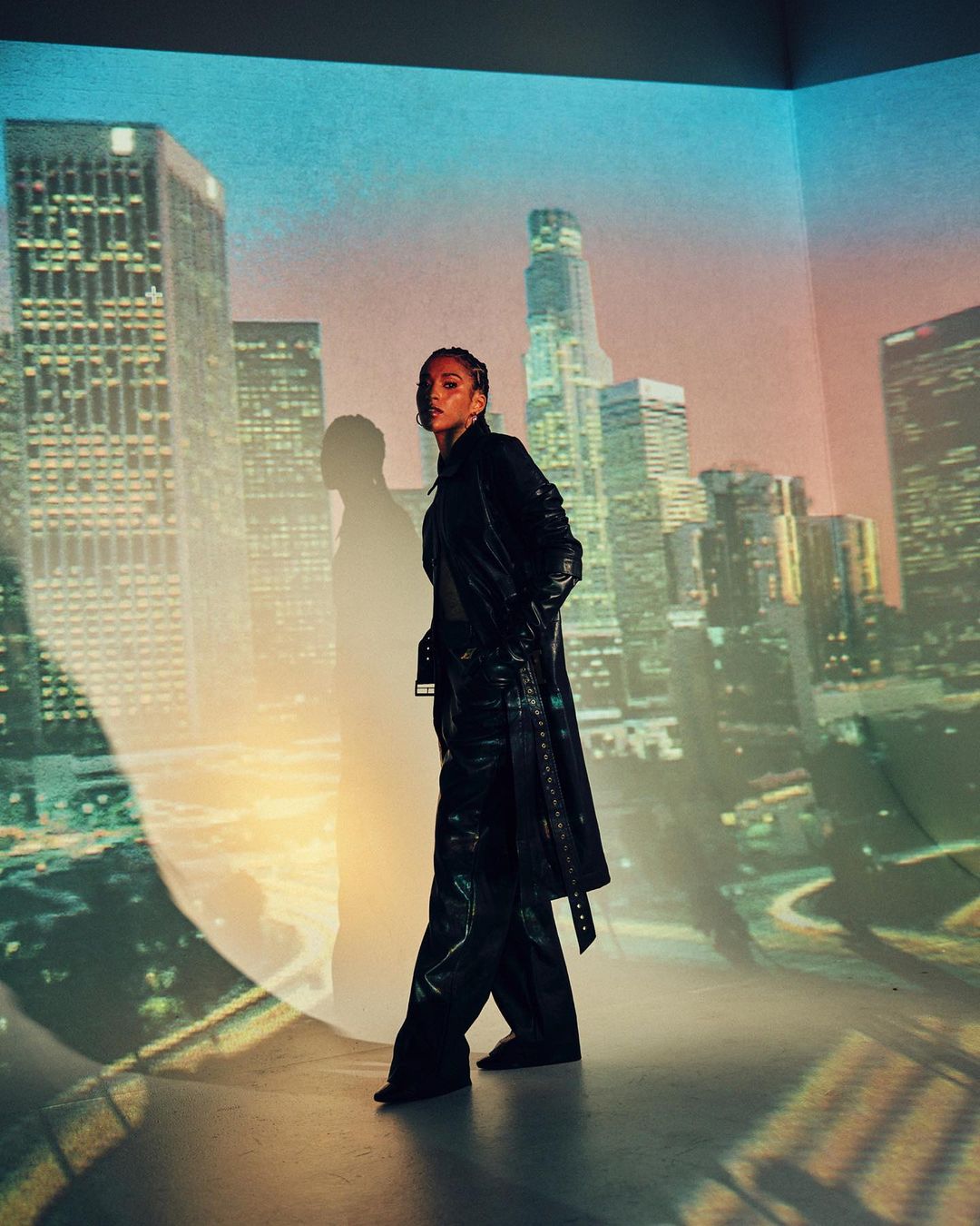 Stylish adult female in black leather outfit poses in an indoor photo studio with a backdrop of projected urban cityscape. 