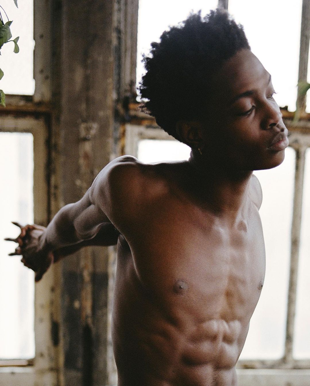 Athletic Black man poses shirtless with outstretched arms in a sunlit, minimalist setting with worn windows and natural light.