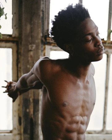 Athletic Black man poses shirtless with outstretched arms in a sunlit, minimalist setting with worn windows and natural light.
