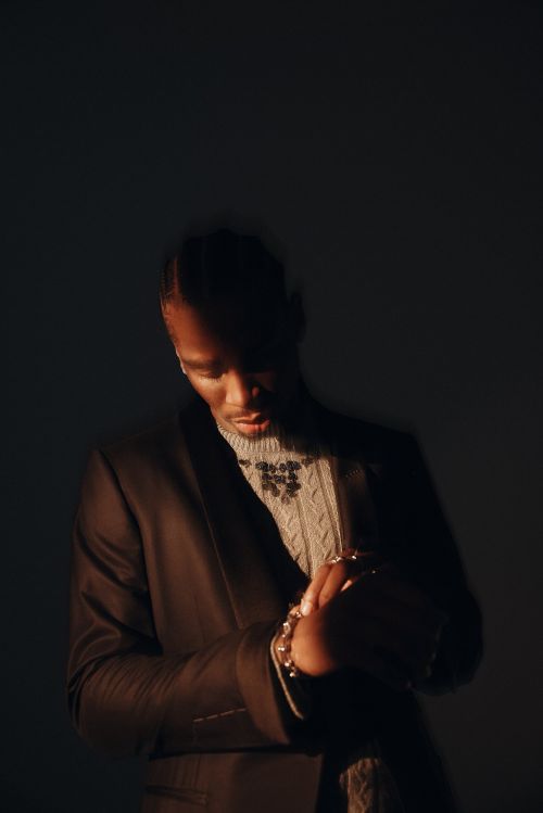 A man in a suit and sweater poses in a studio with dark lighting, showcasing fashion and style with a contemplative expression.