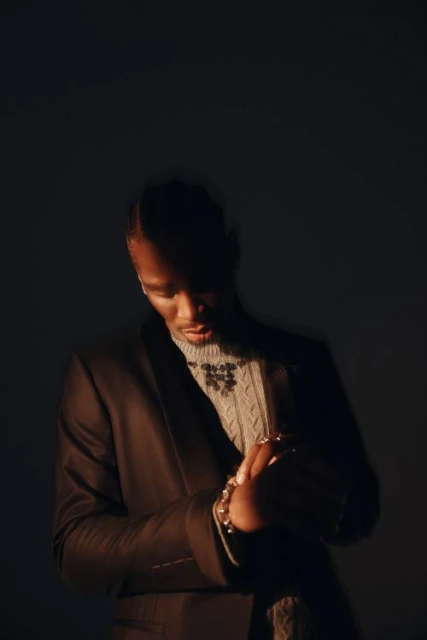 A man in a suit and sweater poses in a studio with dark lighting, showcasing fashion and style with a contemplative expression.